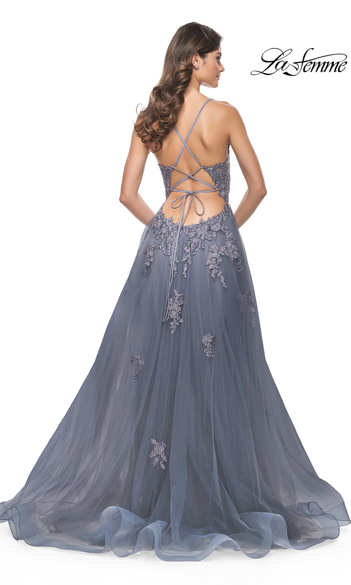 A woman stands facing away in the La Femme Plunging V-Neck Long Prom Ball Gown 31472, a slate blue A-line dress featuring lace appliqué details and a crisscross open back.